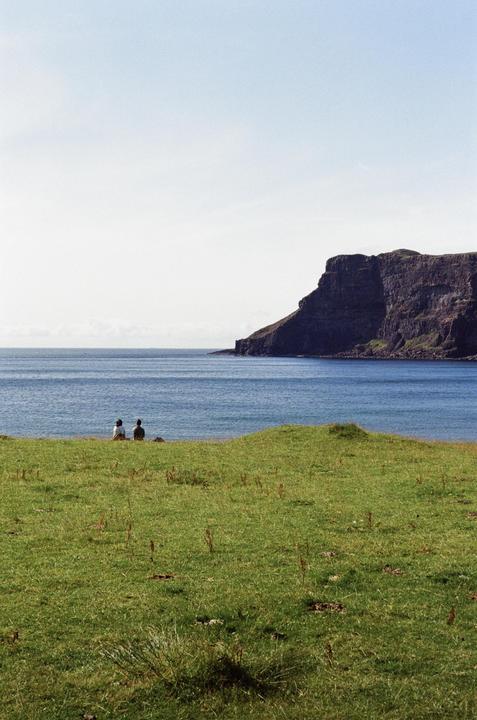 Two people sit on a grassy hill overlooking a body of water and a large cliff.