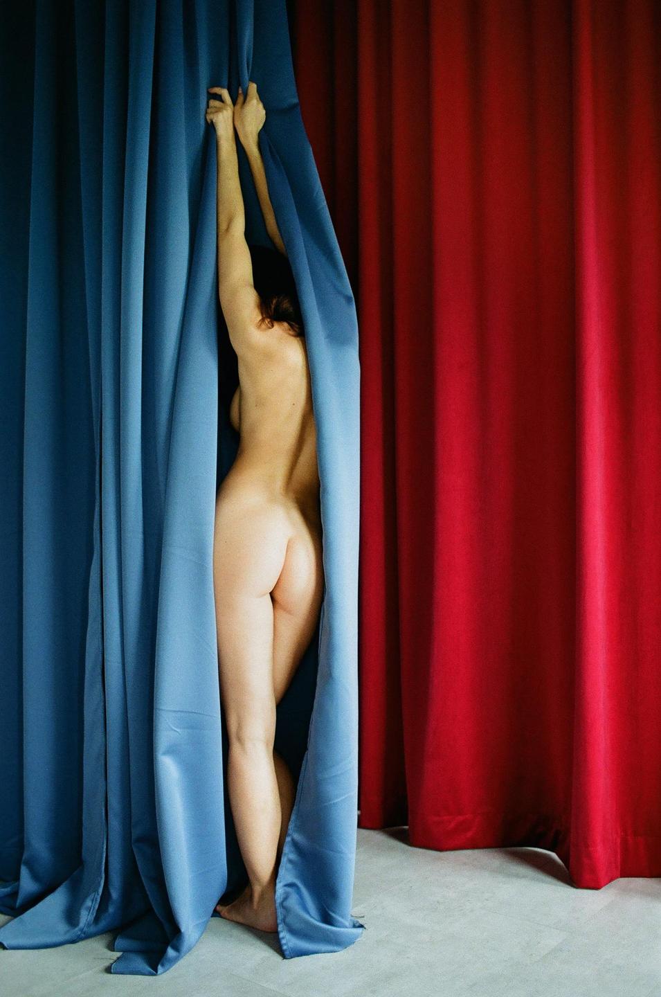 A nude woman stands between blue and red curtains, reaching up to hold the blue curtain.