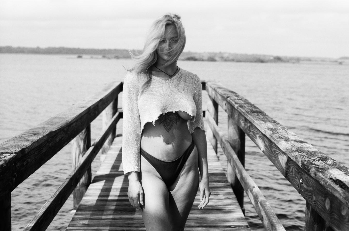 A woman with windblown hair stands on a wooden pier over water.