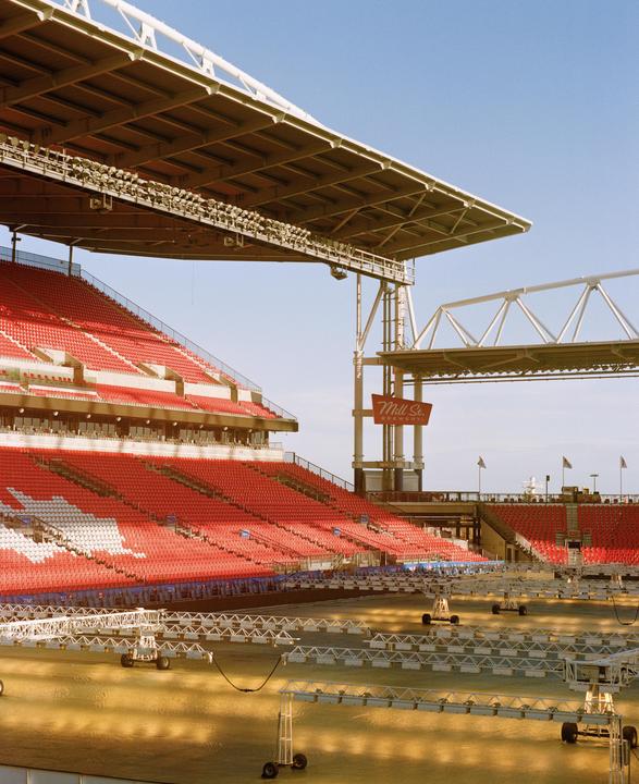 A stadium with red and white seats is visible under a clear sky, with grow lights on the field.
