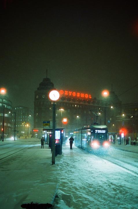 A tram approaches a snowy station at night, with a building displaying a neon sign in the background.