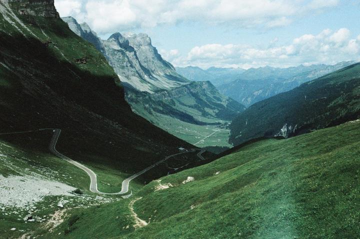 A winding road descends through a green mountain valley under a cloudy sky.