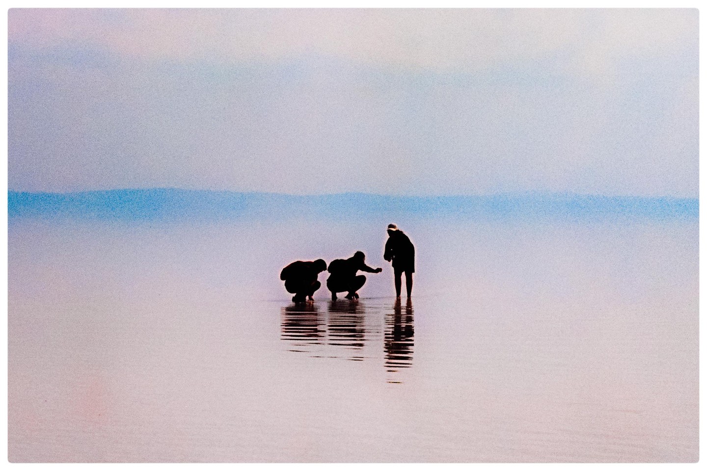 Three people are silhouetted against a hazy sky and water, with two crouching and one standing.