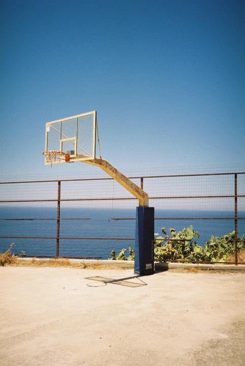 A basketball hoop stands on a court overlooking the ocean, with a fence and cacti in the midground.