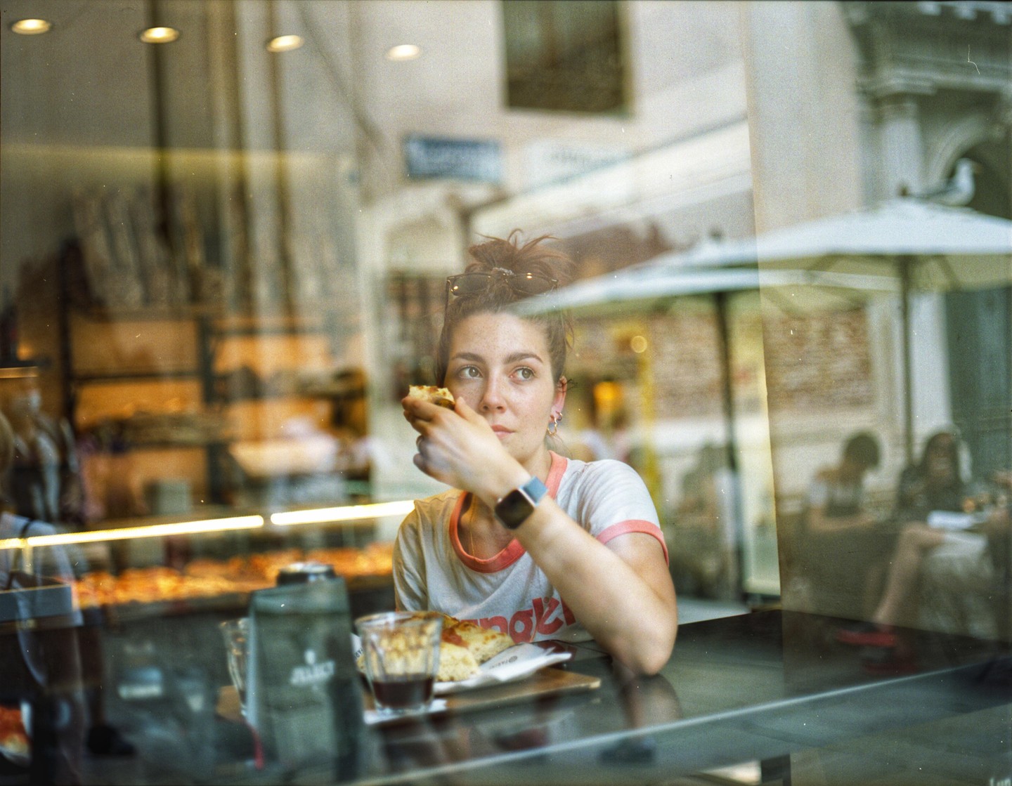 A woman sits at a table inside a cafe, eating a pastry and looking out a window.