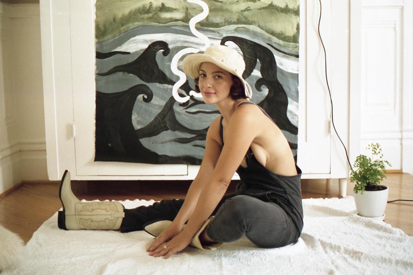 A woman wearing a hat and cowboy boots sits on a white rug in front of a painted cabinet.