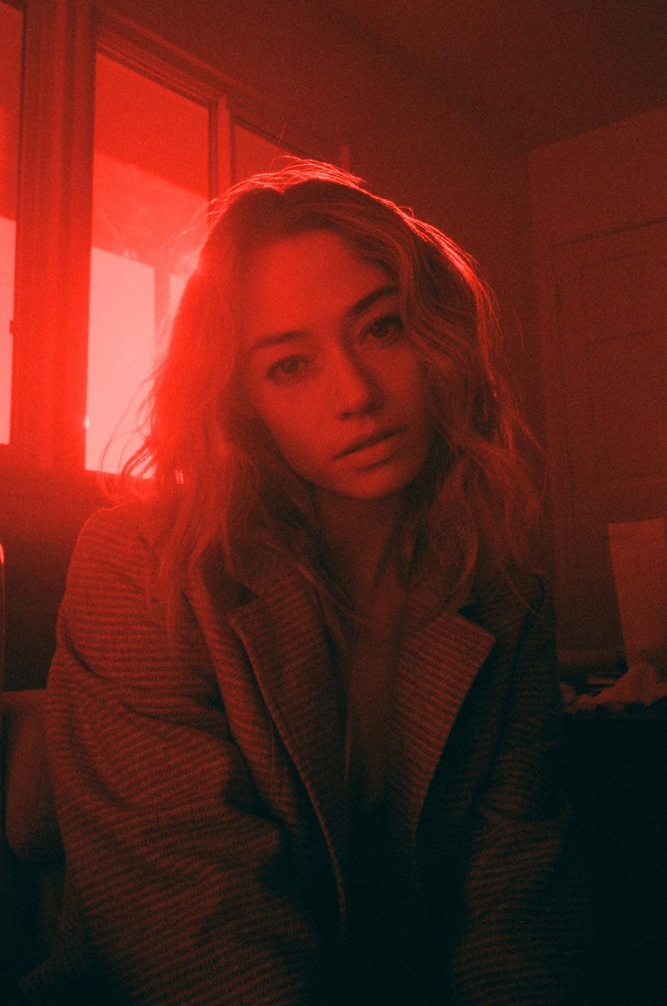 A woman with wavy hair wears a blazer and looks directly at the viewer in a room lit with red light.