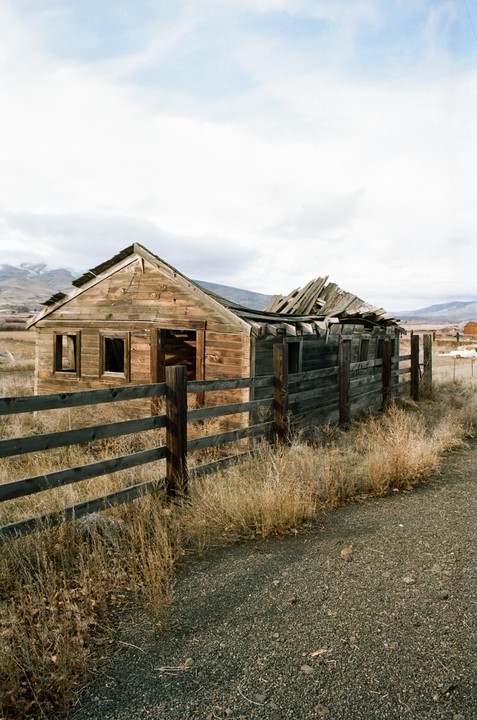 An old wooden shack with a collapsed roof stands behind a wooden fence and dry grass.