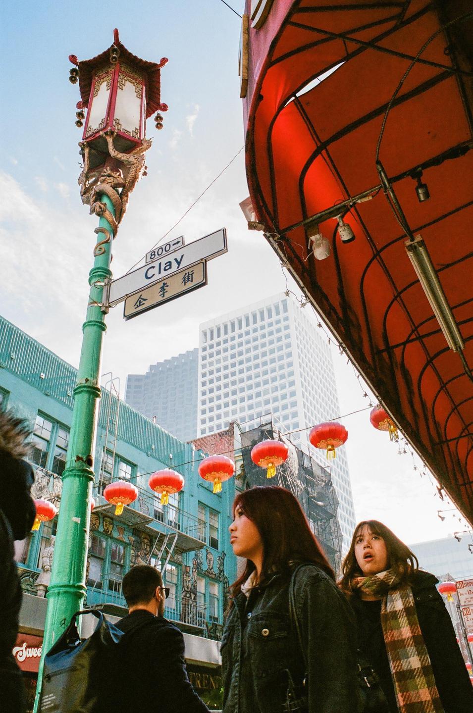 Two women and a man walk along a street in Chinatown, San Francisco, adorned with red lanterns and a decorative street lamp.