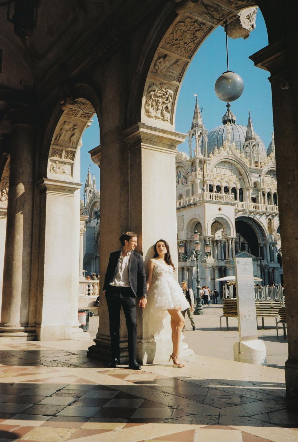 A couple stands under an archway with a large ornate building in the background.