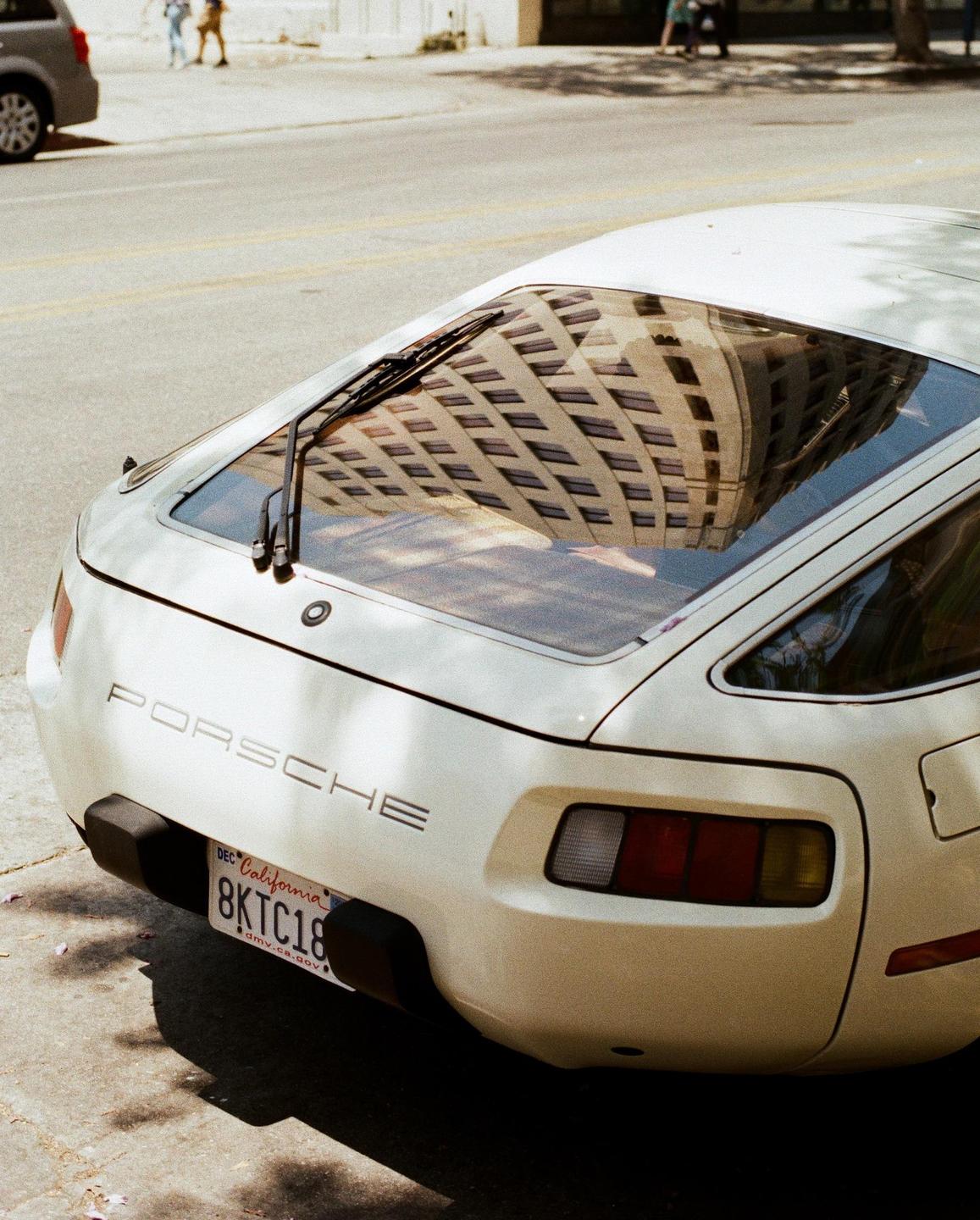 The back of a white Porsche is parked on a street with a building reflected in its rear window.