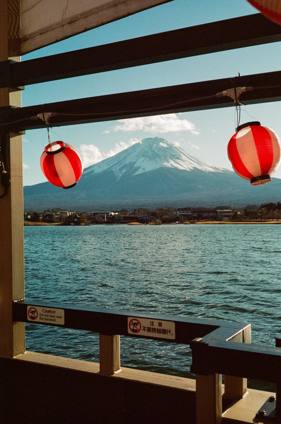 Two red and white lanterns hang in front of a snow-capped mountain and a body of water.