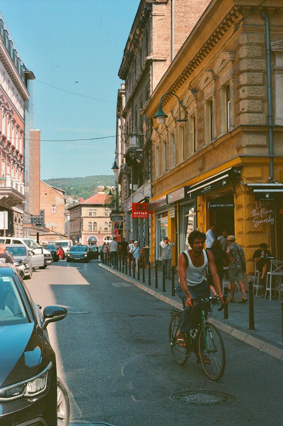 A man rides a bicycle down a street lined with buildings and parked cars, with people walking on the sidewalk.