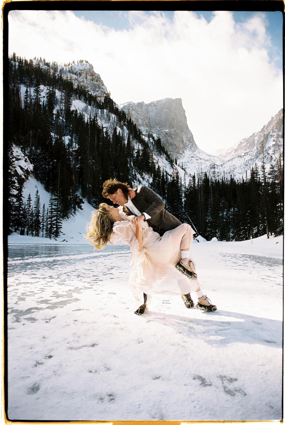 A couple dances on a frozen lake surrounded by snow-covered mountains and pine trees.