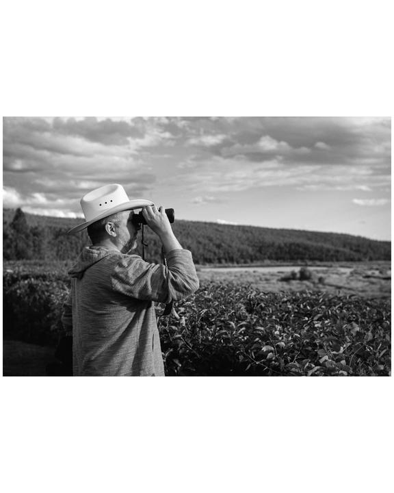 A man wearing a cowboy hat looks through binoculars at a distant landscape.