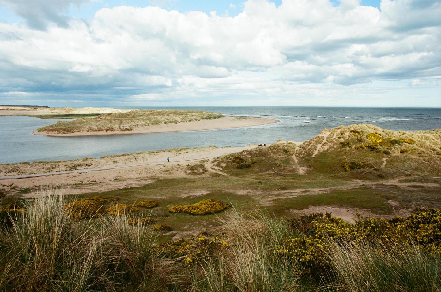 A wide view shows a river meeting the sea, with sand dunes and green vegetation under a cloudy sky.