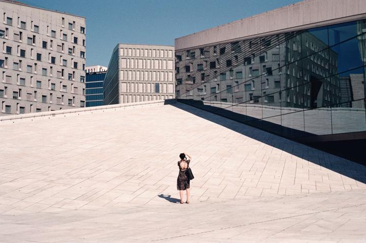 A woman stands on a large, light-colored paved surface, looking towards modern buildings with reflective windows.
