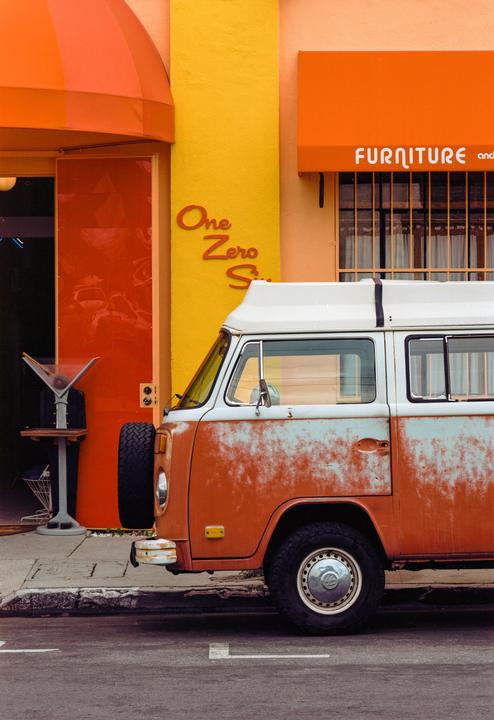 A rusty orange and white van is parked on a street in front of a building with orange and yellow walls and awnings.