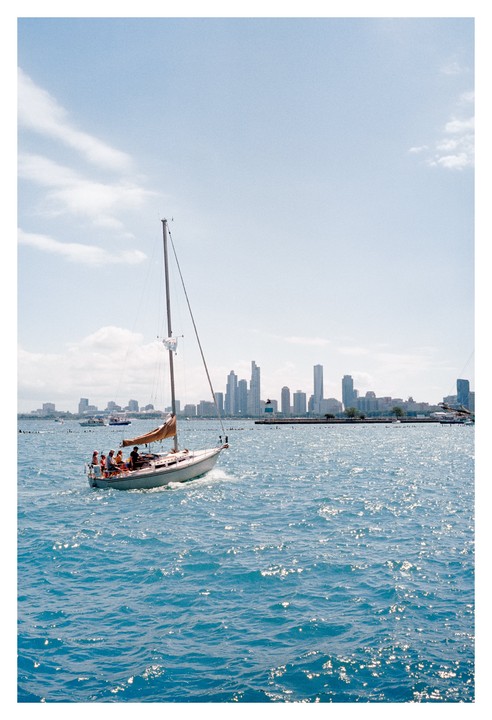 A sailboat with people on board moves across sparkling blue water with a city skyline in the background.