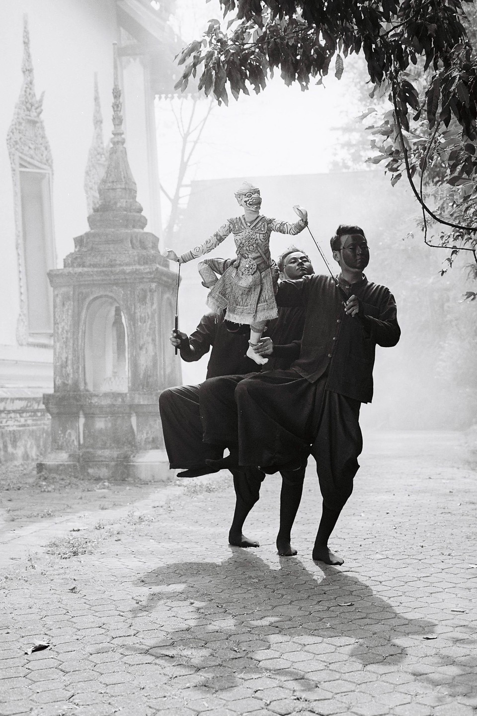 Three men in dark clothing perform a traditional Thai puppet show on a paved path near a temple.