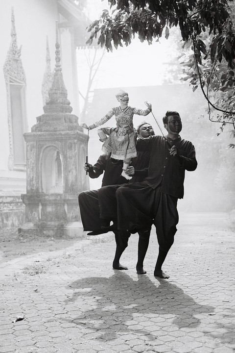 Three men in dark clothing perform a traditional Thai puppet show on a paved path near a temple.