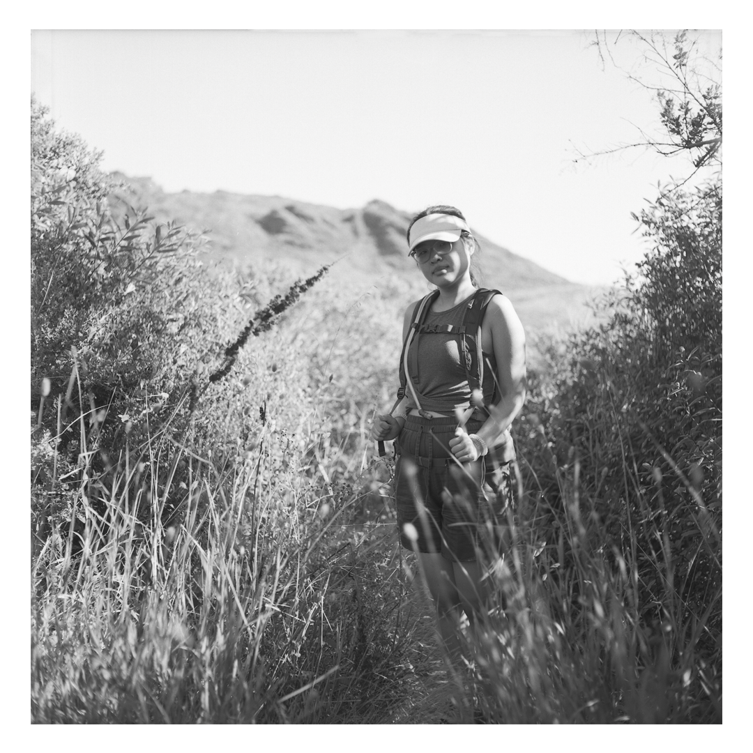 A woman wearing a visor and backpack stands in a field of tall grass with a mountain in the background.
