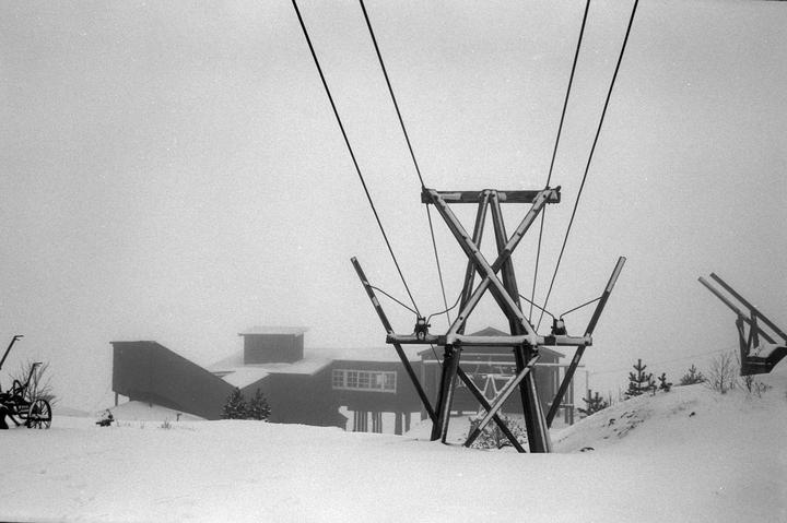 A ski lift tower stands in a snow-covered landscape with a building in the background.