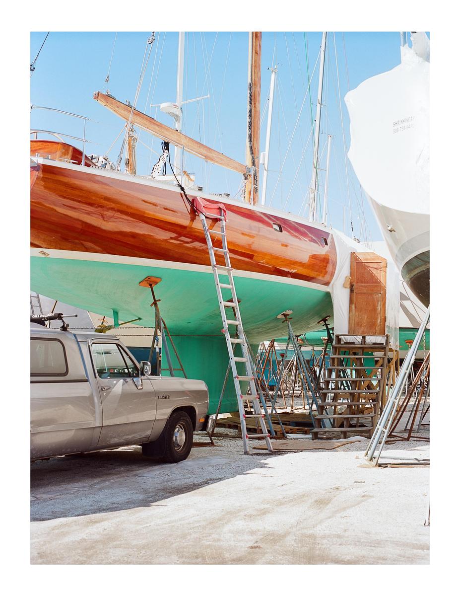 A boat is propped up on stands next to a truck and a ladder in a boatyard.