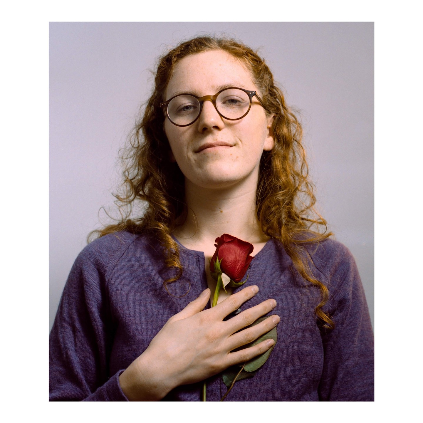 A woman with curly red hair and glasses holds a single red rose to her chest.