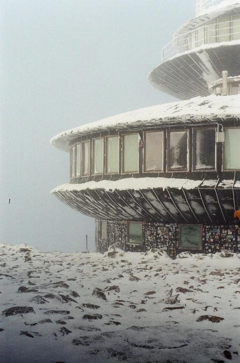 A snow-covered building with many windows stands on a rocky, snowy landscape under a hazy sky.