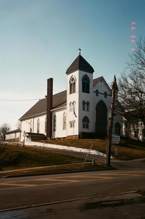 A white church with a tall steeple and a cross on top stands on a grassy hill next to a road.