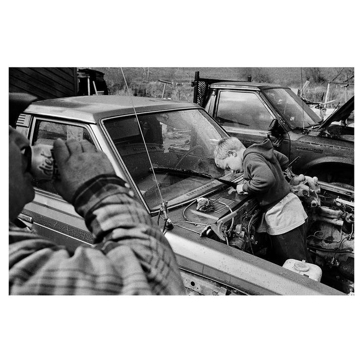 A man drinks from a can while a boy works on a car engine in a black and white outdoor scene.