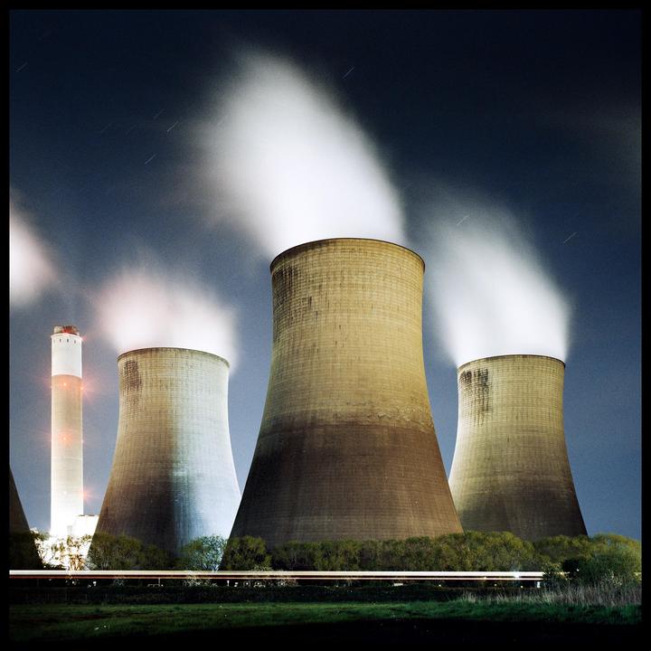 Three cooling towers and a tall smokestack emit steam into the night sky, with a blurred train passing in the foreground.