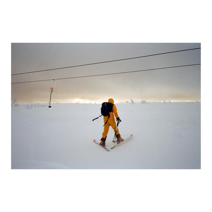 A person in a yellow ski suit skis down a snowy slope under a cloudy sky.