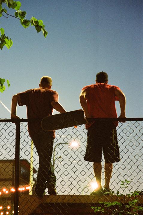 Two men stand with their backs to the viewer, leaning on a chain-link fence with a skateboard between them, under a clear sky with bright lights in the background.