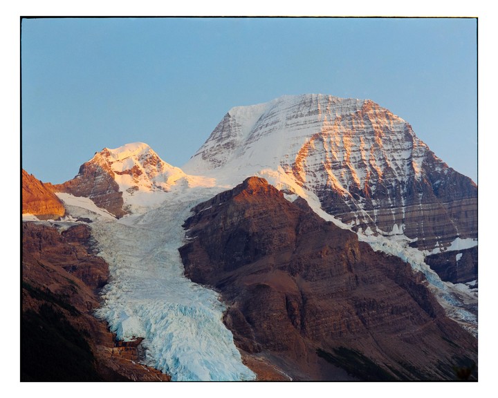 A snow-capped mountain with a glacier is illuminated by alpenglow under a clear sky.