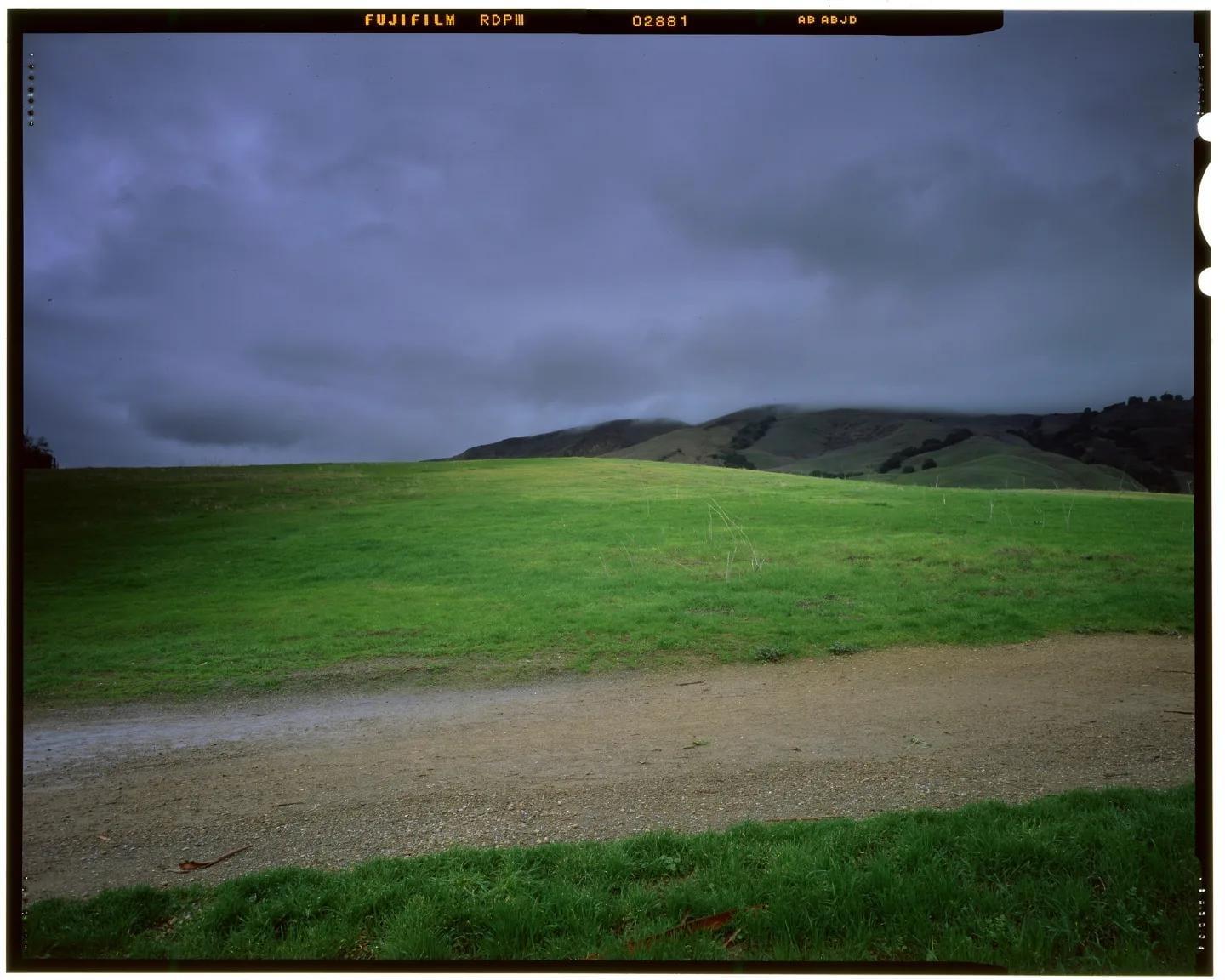 A dirt path leads through a green field towards hills partially obscured by fog under a cloudy sky.