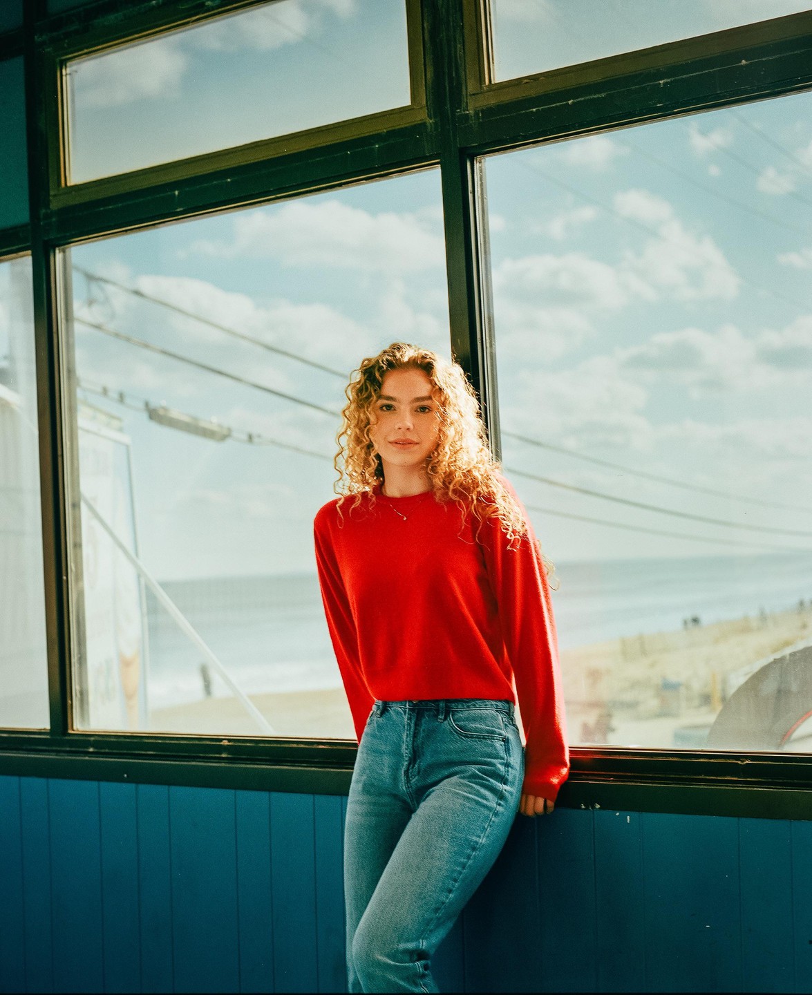 A woman with curly hair wearing a red sweater and jeans stands in front of a window overlooking a beach.