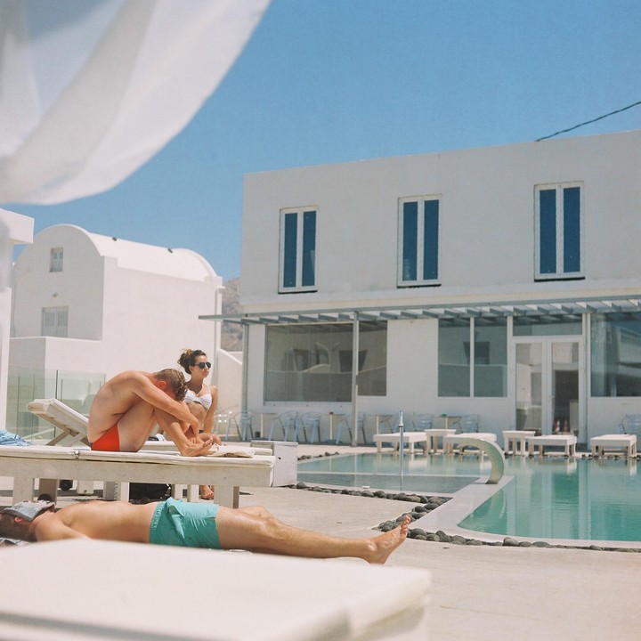 Three people are relaxing by a swimming pool in front of white buildings under a clear blue sky.