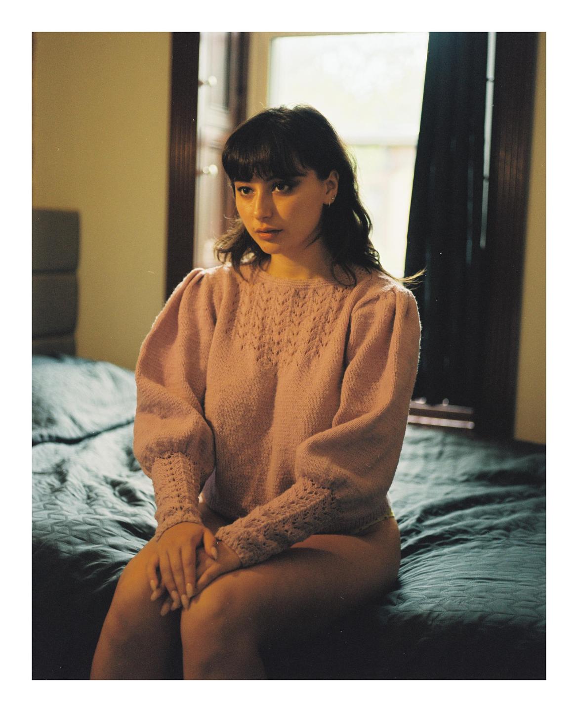 A woman with dark hair and bangs sits on a bed wearing a pink knitted sweater.
