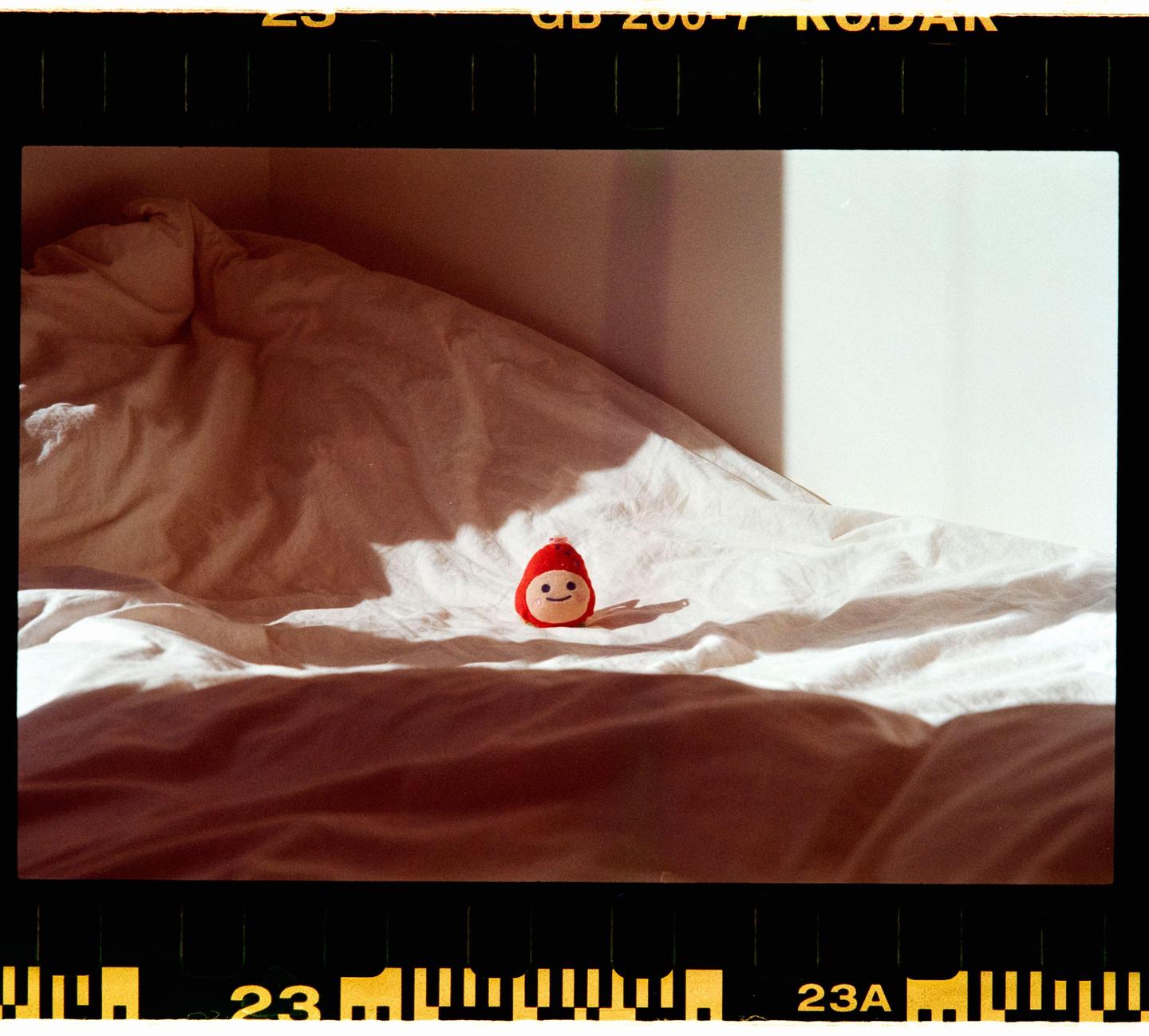 A small red doll with a smiling face sits on a white bedspread in a sunlit room.