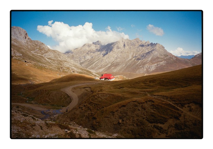 A red-roofed building sits in a valley surrounded by mountains under a blue sky with clouds.