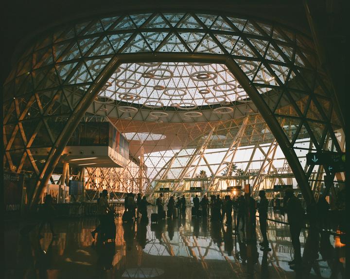 People stand on a reflective floor under a large glass dome in an airport at sunset.