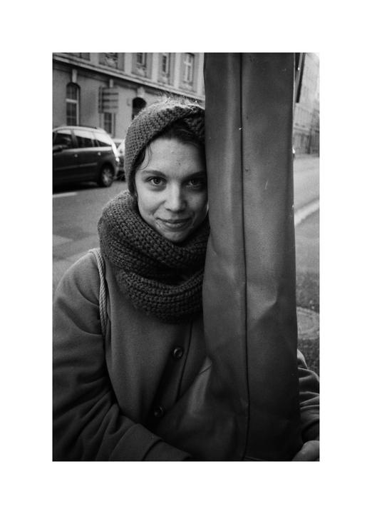 A woman wearing a knitted hat and scarf smiles while standing next to a pole on a city street.