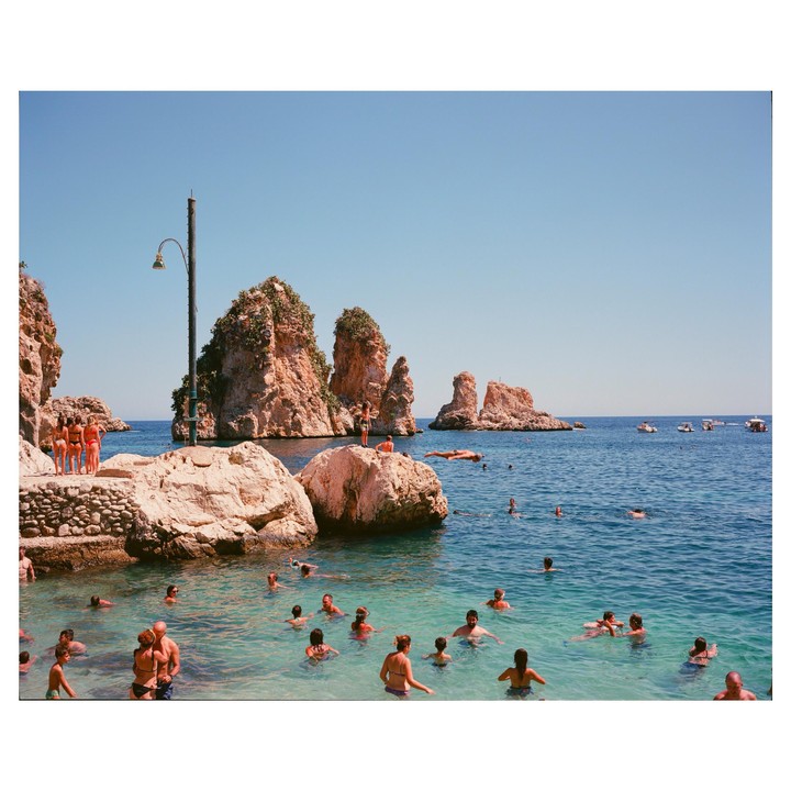 People swim in clear blue water near large rock formations under a bright sky.