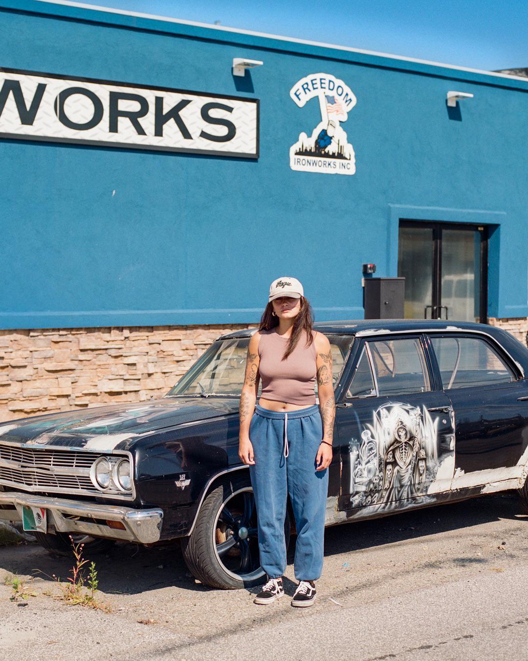 A woman stands in front of a black car with a mural on its side, parked in front of a blue building.