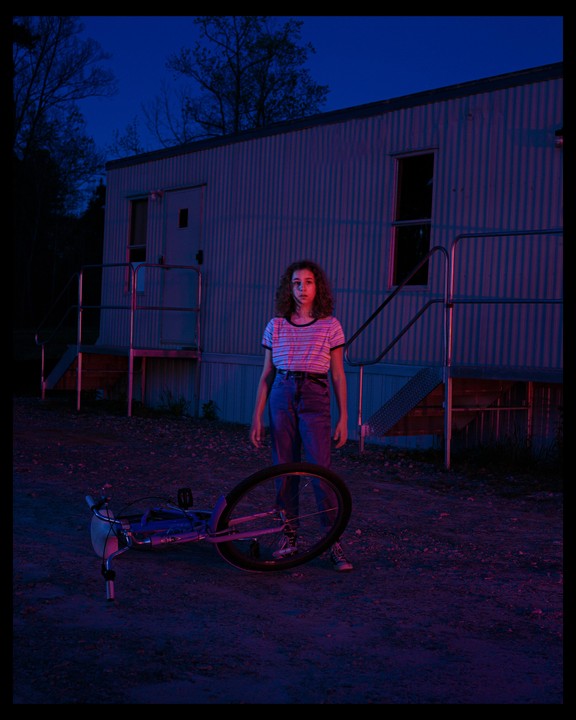 A young woman stands in front of a trailer at night, with a bicycle lying on the ground.