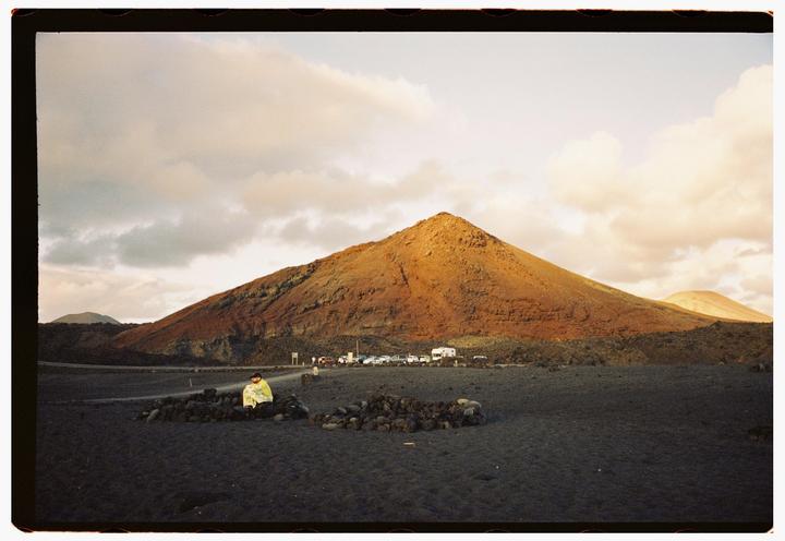 A person sits wrapped in a blanket on a dark, rocky plain with a large, reddish-brown mountain in the background under a cloudy sky.