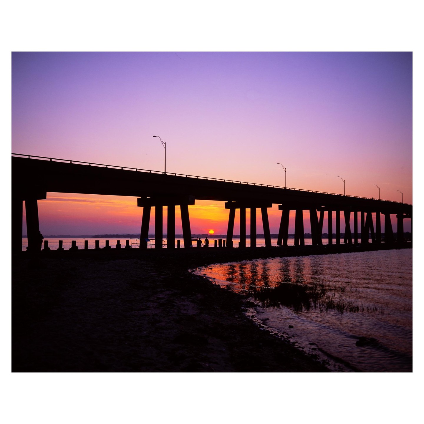 A bridge stretches over water with the sun setting beneath its arches, casting a vibrant glow on the water.