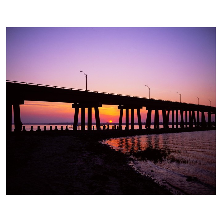 A bridge stretches over water with the sun setting beneath its arches, casting a vibrant glow on the water.